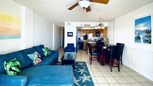 A living room with a blue sofa, a dining table, and a kitchen in the background. - SunSwept 505 by ALBVR (Orange Beach)