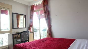 A bedroom with a bed, a desk, and a chair. There is a window with curtains and a view of the outside.