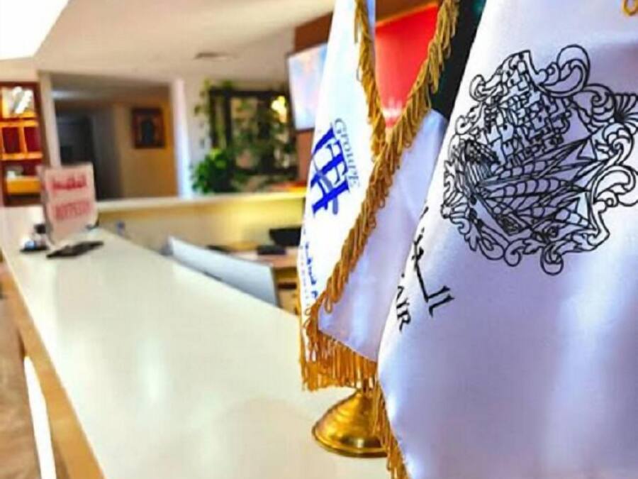 Un área de recepción con un mostrador blanco, un cartel que dice "Bienvenido" y dos banderas con emblemas.