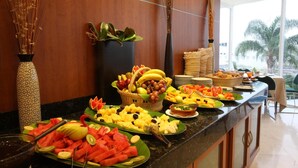A buffet with various fruits and vegetables, a vase with dried plants, and a view of palm trees outside. - Hotel Mercury Inn (Querétaro)