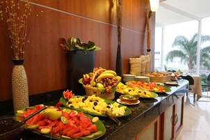 Un bufé con diversas frutas y verduras, un jarrón con plantas secas y vistas a palmeras afuera.