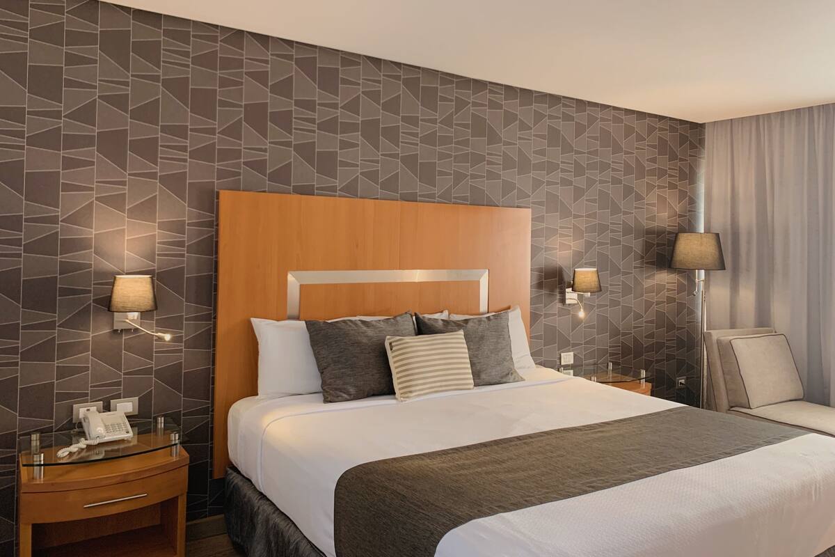 A modern hotel room with a geometric-patterned wall, a wooden headboard, a large bed with multiple pillows, bedside lamps, a bedside table with a phone, and a seating area with a chair and a lamp.