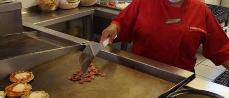 Uma cozinha com uma chapa, uma pessoa cozinhando e diversos alimentos.
