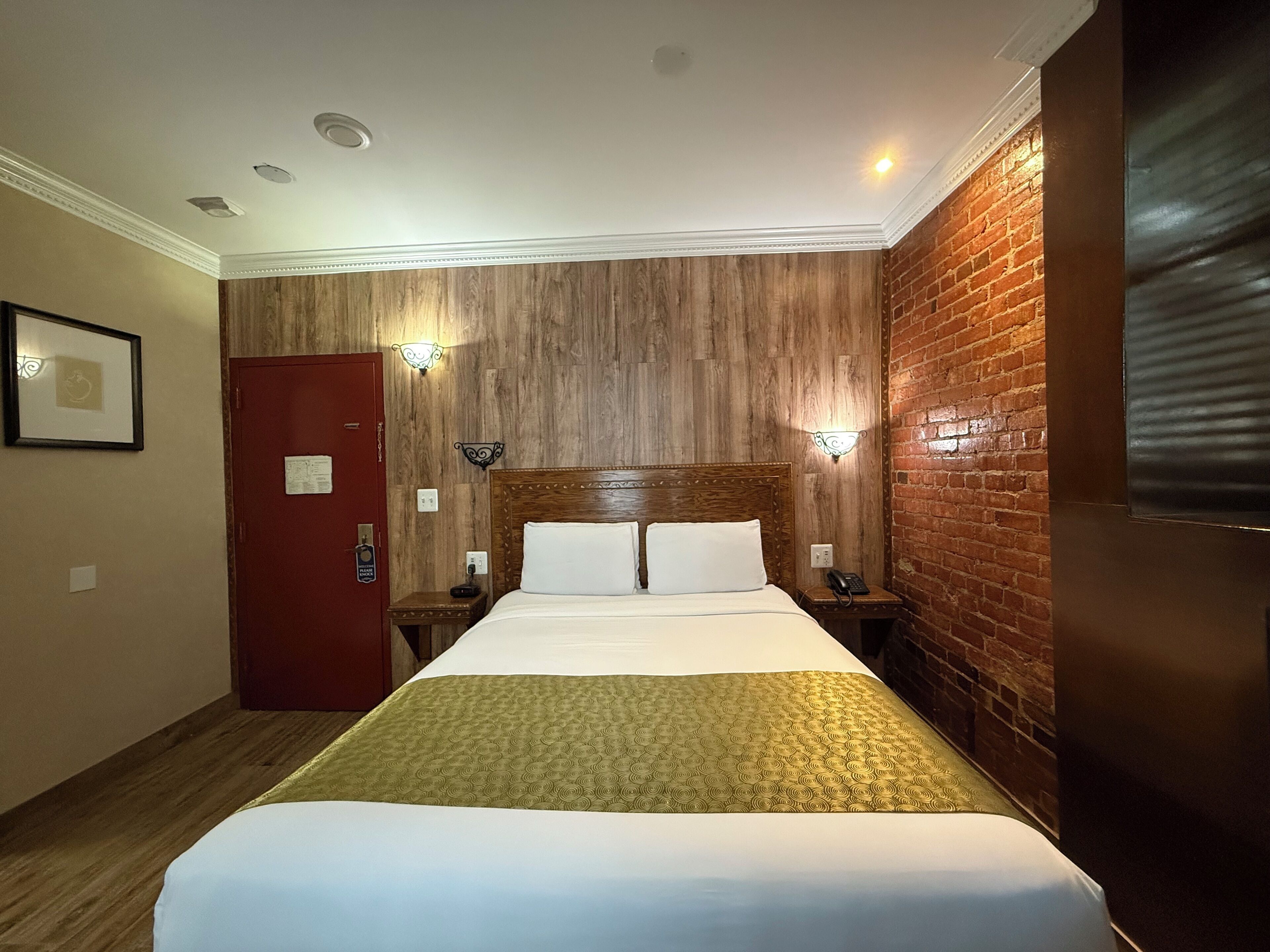 Une chambre d’hôtel avec une tête de lit en bois, un lit recouvert de draps blancs, deux tables de chevet avec des lampes, une porte rouge et un mur en briques.