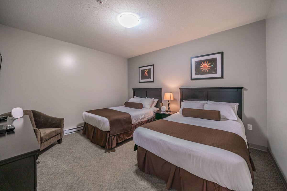 A hotel room with two beds, a desk, a chair, and a lamp.