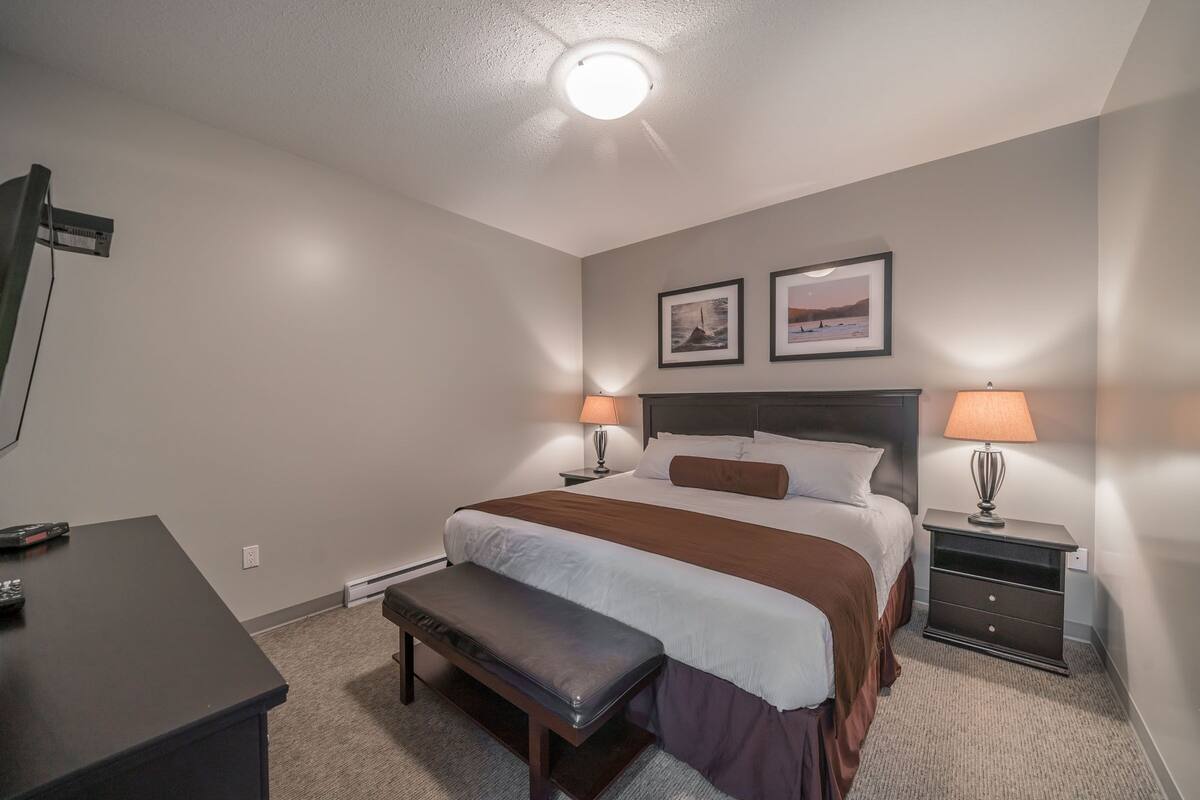 A hotel room with a bed, bedside tables with lamps, a bench, a desk, and a ceiling light.