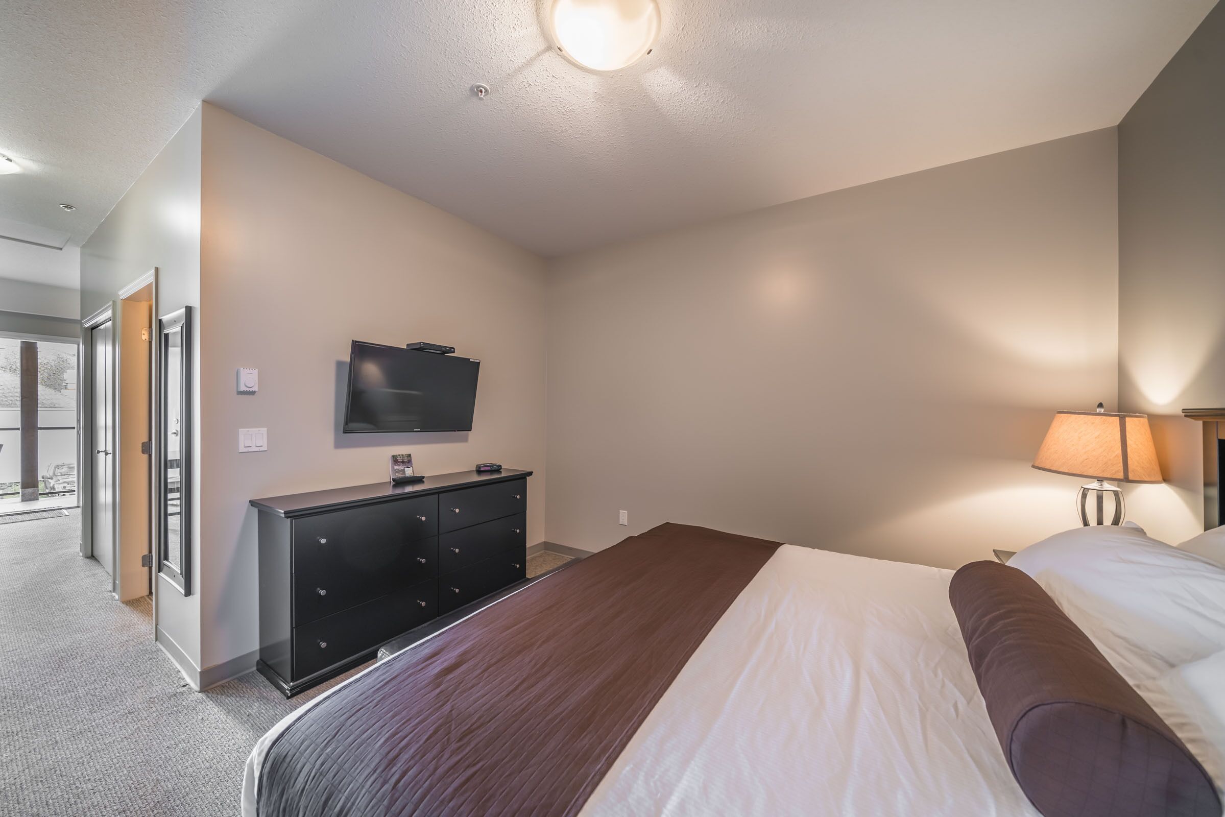 Habitación de hotel con una cama grande, televisión fijada a la pared, cómoda y lámpara.