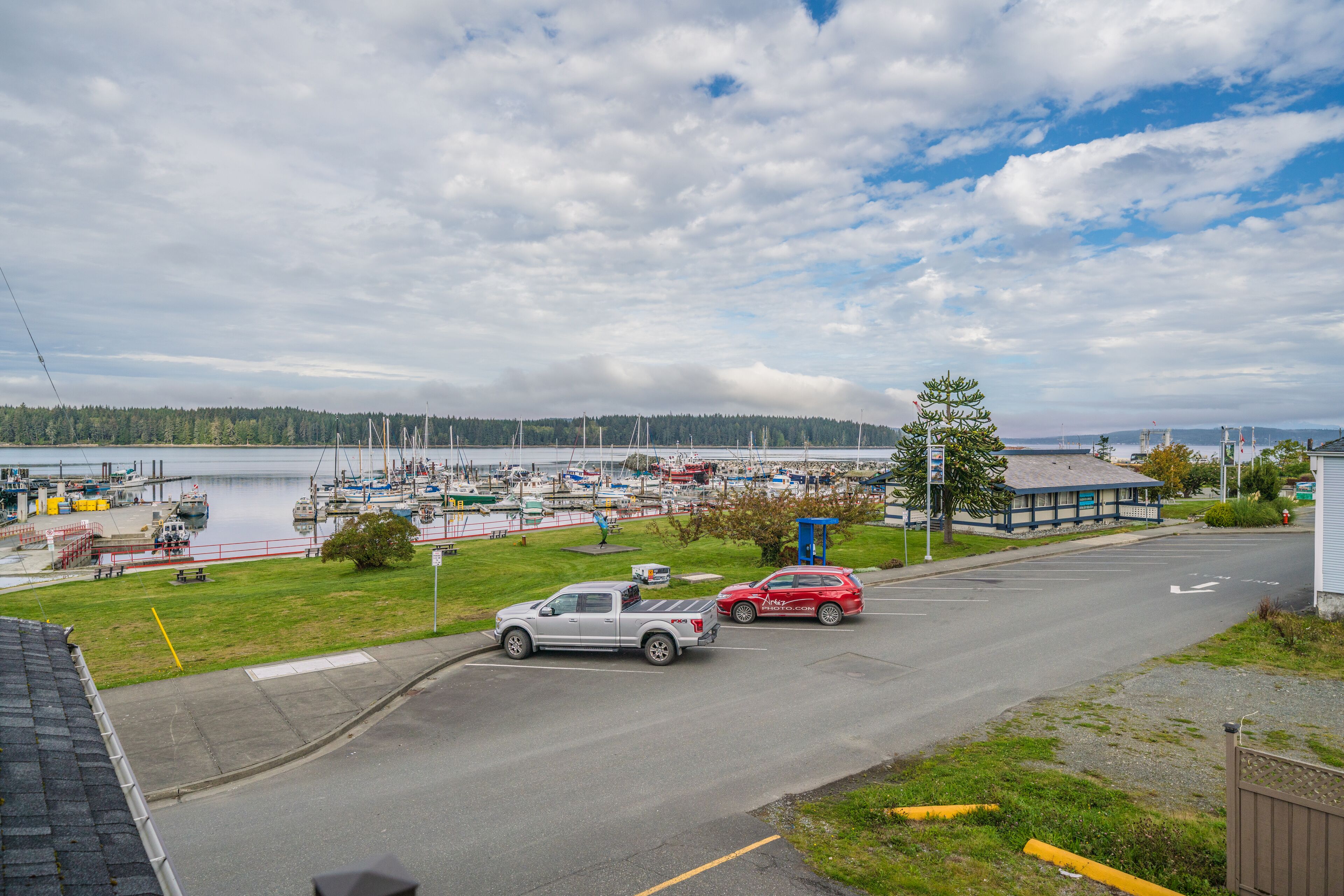 Un port de plaisance avec des bateaux, une aire de stationnement avec un camion et une voiture, et un bâtiment avec un toit bleu.