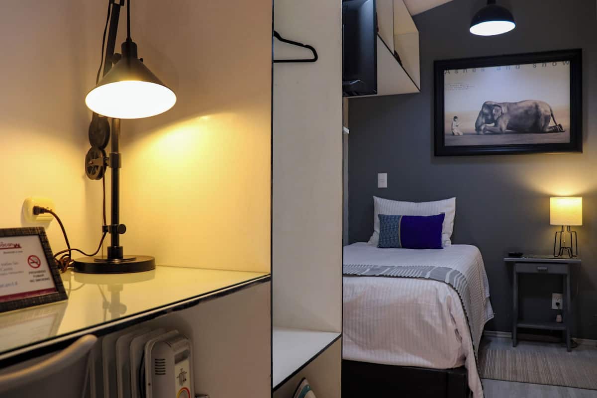 A hotel room with a desk lamp, a framed picture on the wall, a bed with a blue pillow, and a bedside table with a lamp.