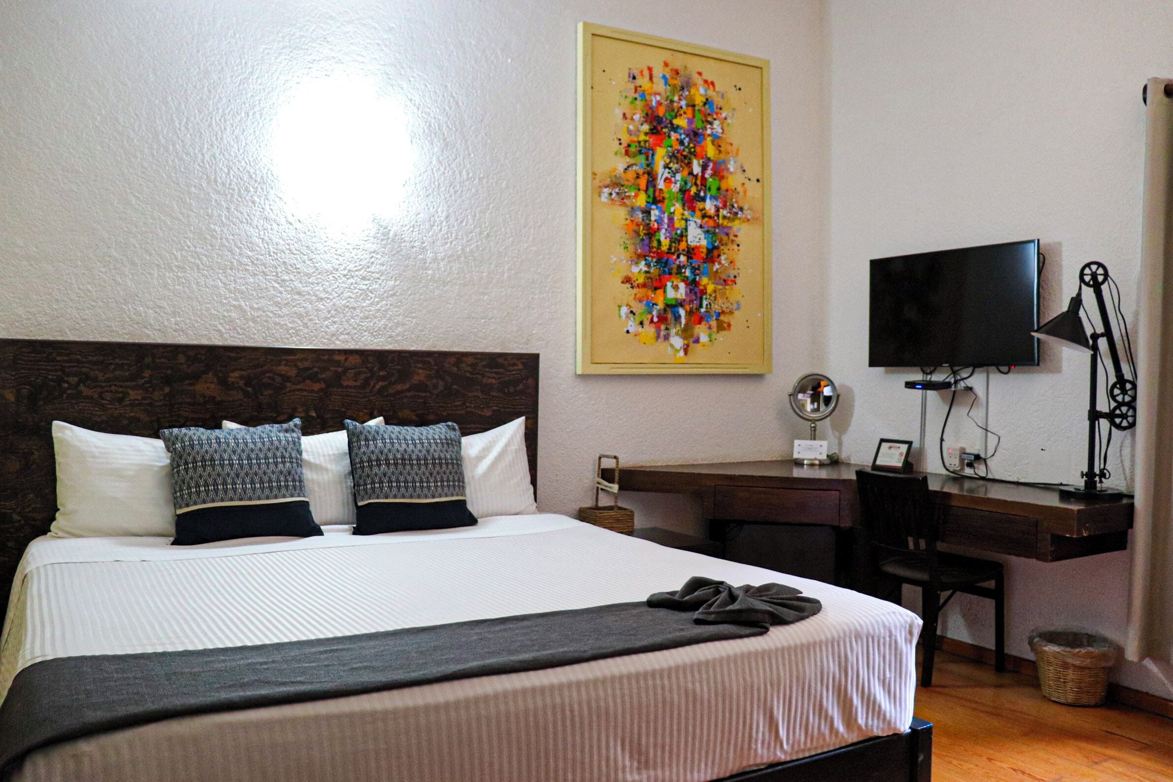 A neatly made bed with a white and black bedspread, a wooden headboard, and two pillows. A colorful abstract painting is on the wall above the bed. A desk with a computer and a TV is to the right of the bed.