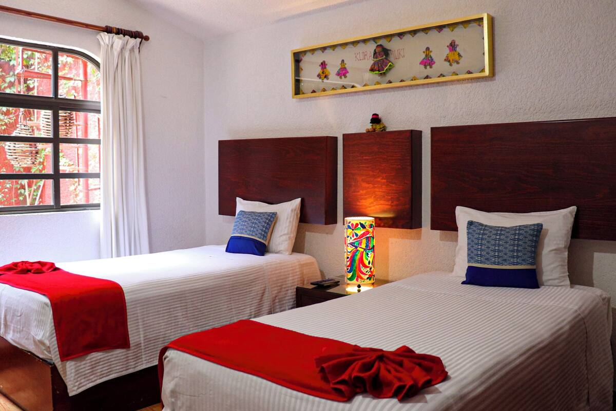 Two single beds with white bedding and red throw blankets, a nightstand with a lamp, and a window with a curtain.
