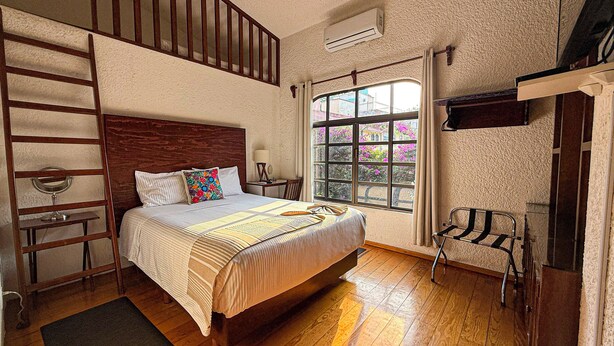 Un dormitorio con cama de madera, una ventana con vistas a edificios y flores, una escalera y un equipo de aire acondicionado.