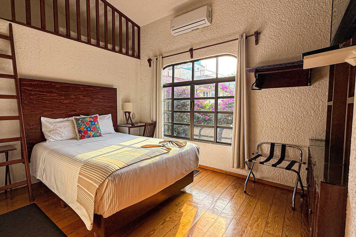 A bedroom with a wooden bed, a window with a view of buildings and flowers, a ladder, and an air conditioning unit.