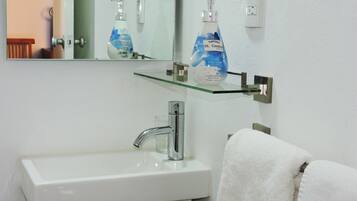 Un baño con un lavabo blanco, un espejo y una estantería de vidrio con artículos de aseo.