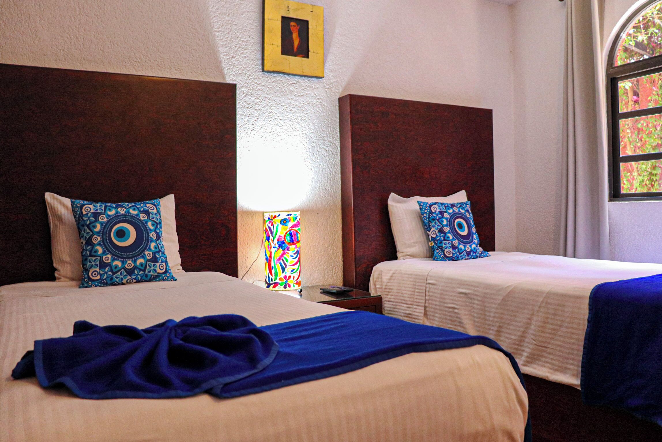 A hotel room with two beds, each with a patterned pillow and a blue blanket.