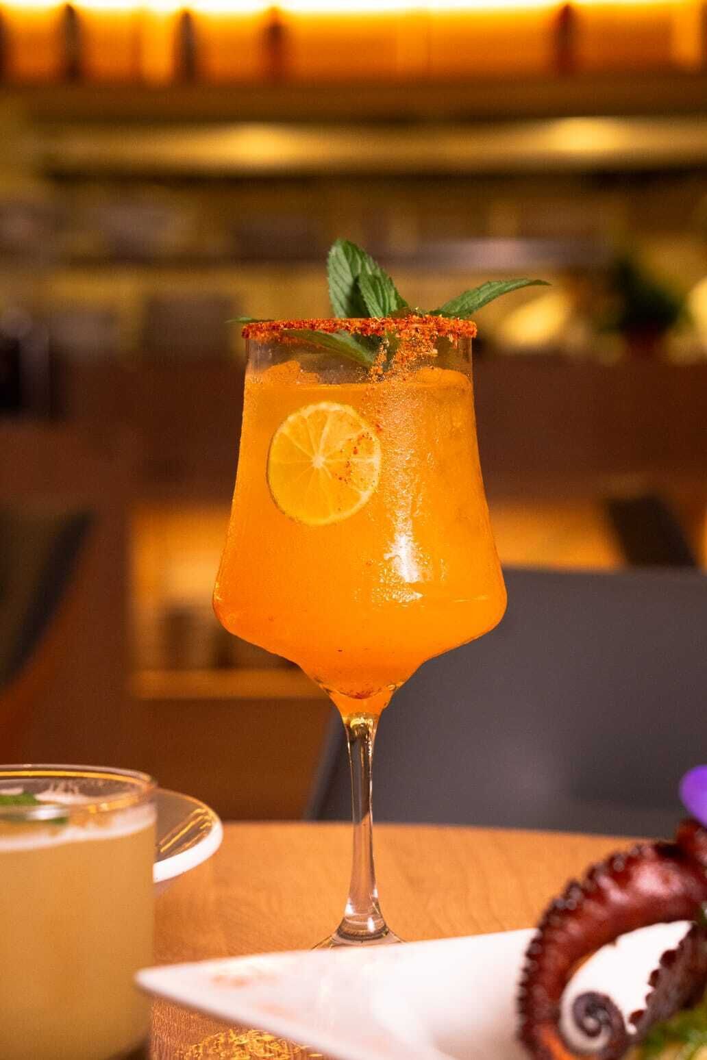 A cocktail in a glass with a lemon slice and mint garnish, placed on a wooden table with a plate of octopus in the background.