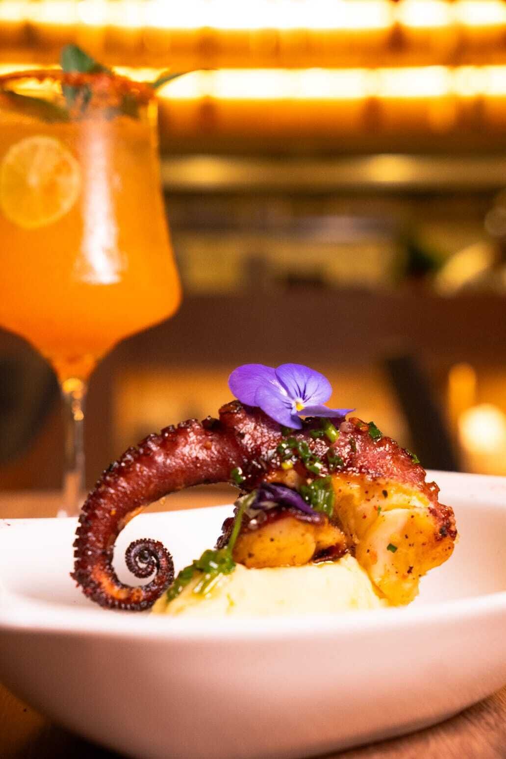 Een bord met octopus en een paarse bloem als garnering, samen met een glas oranje drank.