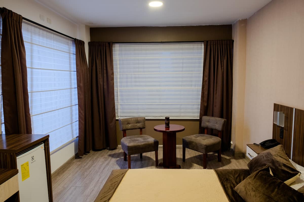 A hotel room with a bed, two armchairs, a small table, a desk with a computer, a telephone, and a window with blinds.