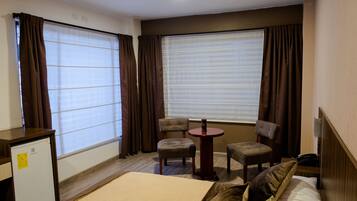 A hotel room with a bed, two chairs, a small table, a desk, and a window with curtains.