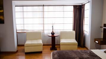 A hotel room with a large window, two armchairs, a small round table with a vase, and a bed with a brown blanket.