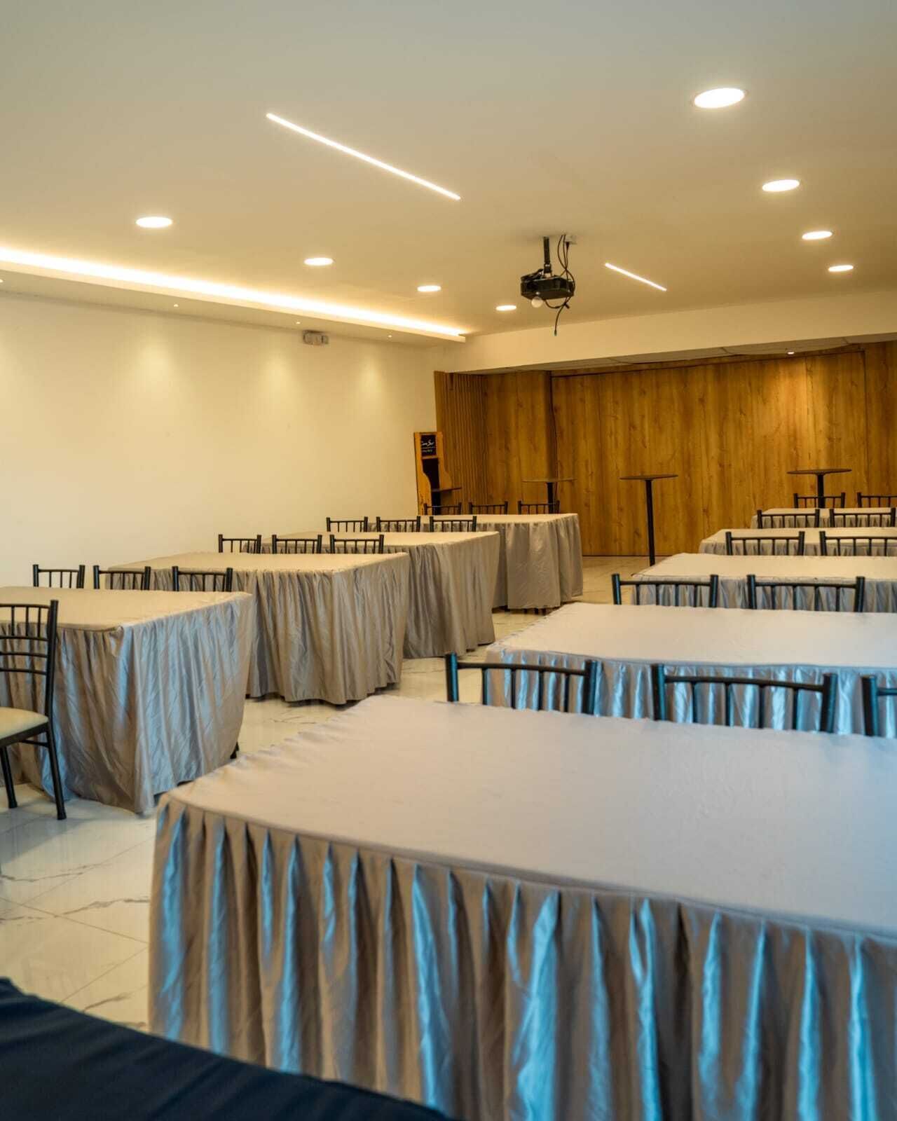 A conference room with multiple round tables, each covered with a beige tablecloth. The room has a wooden panel wall and recessed ceiling lighting.
