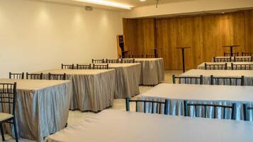 A conference room with multiple round tables, each covered with a beige tablecloth. The room has a wooden panel wall and recessed ceiling lighting.