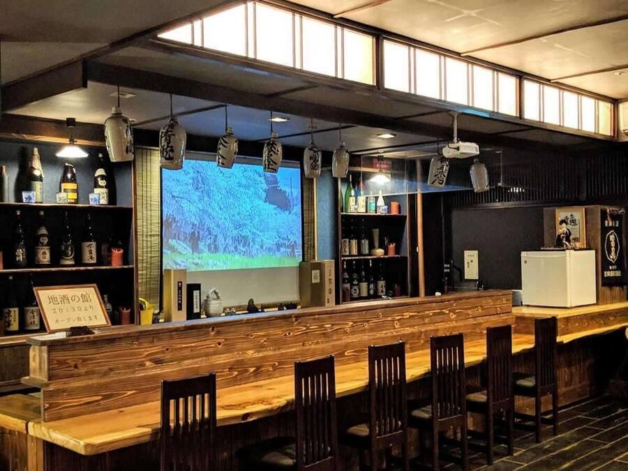 A bar area with wooden bar counter and chairs, shelves with bottles, and a projection screen displaying a landscape.