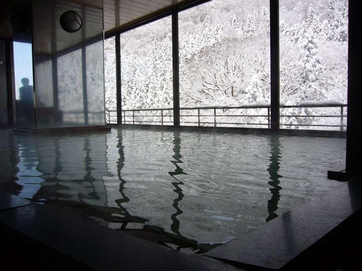 A large indoor swimming pool with clear reflections on the water surface, surrounded by large windows offering views of a snowy landscape.