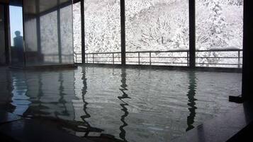 A large indoor swimming pool with clear reflections on the water surface, surrounded by large windows offering views of a snowy landscape.