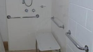 Un baño pequeño con azulejos, ducha, barras de apoyo y una ventana.