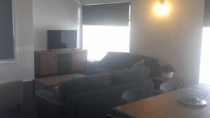 Un espacio habitable compacto con sofá, una mesita, televisor sobre un mueble y ventana con persianas.