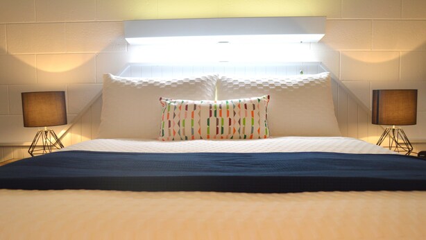 Una habitación de hotel con una cama grande, ropa de cama blanca, una almohada colorida, dos lámparas de noche y un cabecero con iluminación integrada.