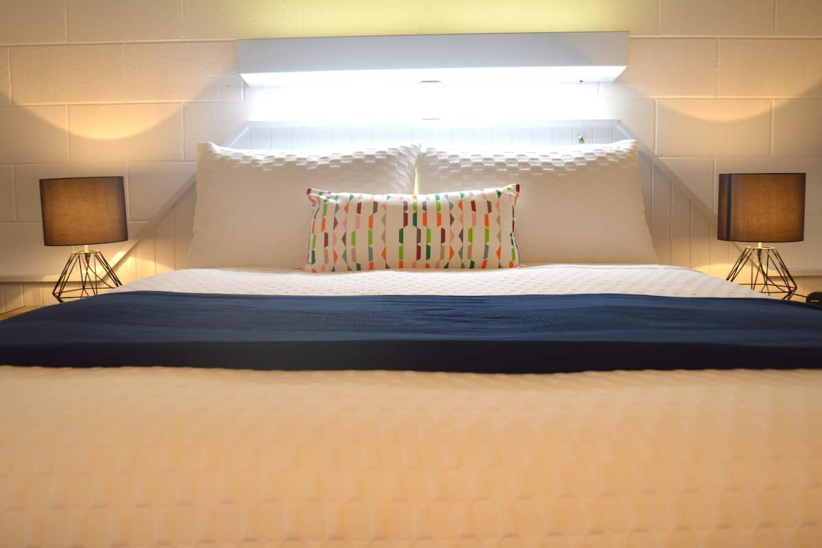 Una habitación de hotel con una cama grande, ropa de cama blanca, una almohada colorida, dos lámparas de noche y un cabecero con iluminación integrada.