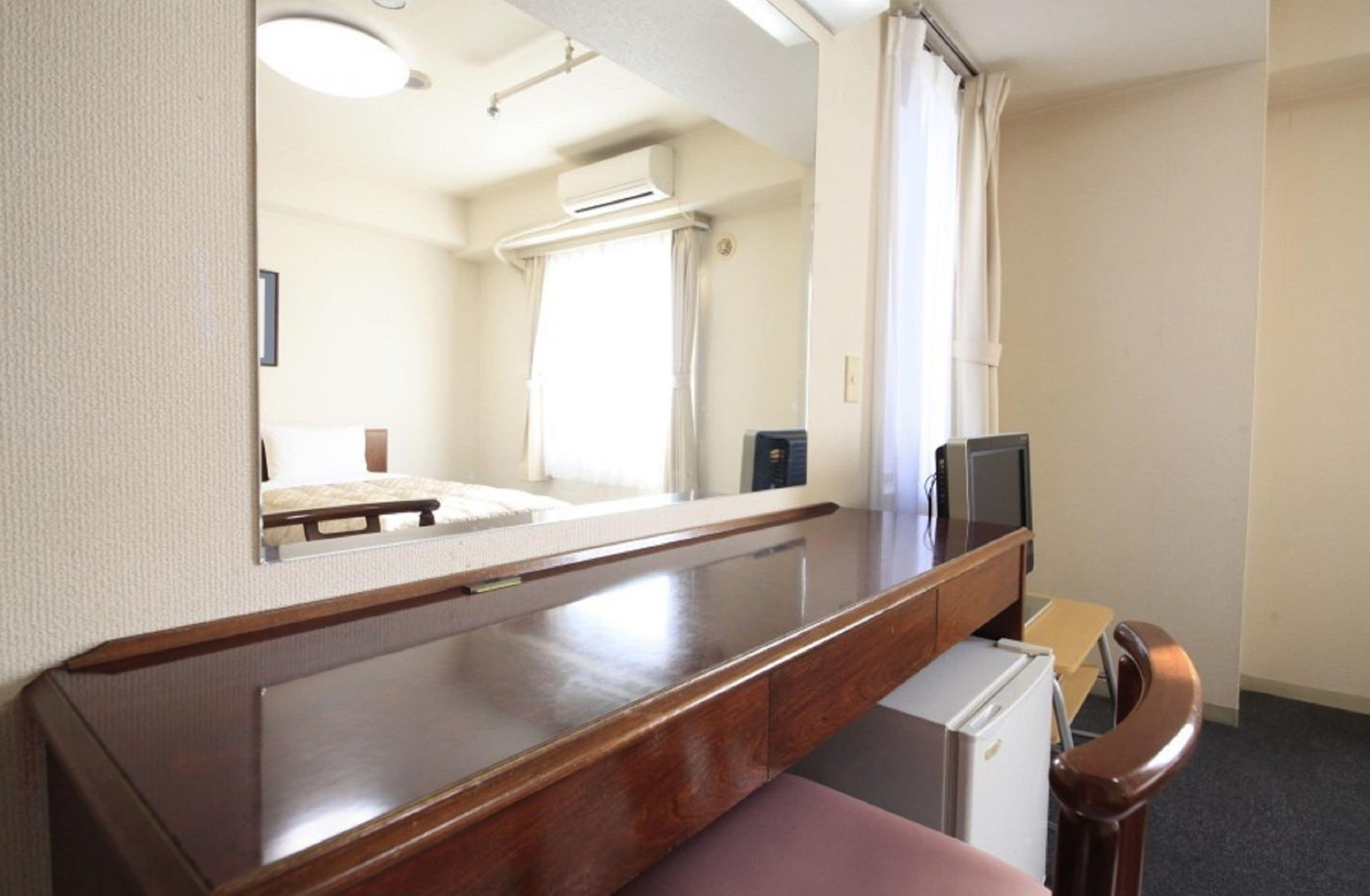 A compact hotel room with a wooden desk, a small refrigerator, a television, and a mirror.