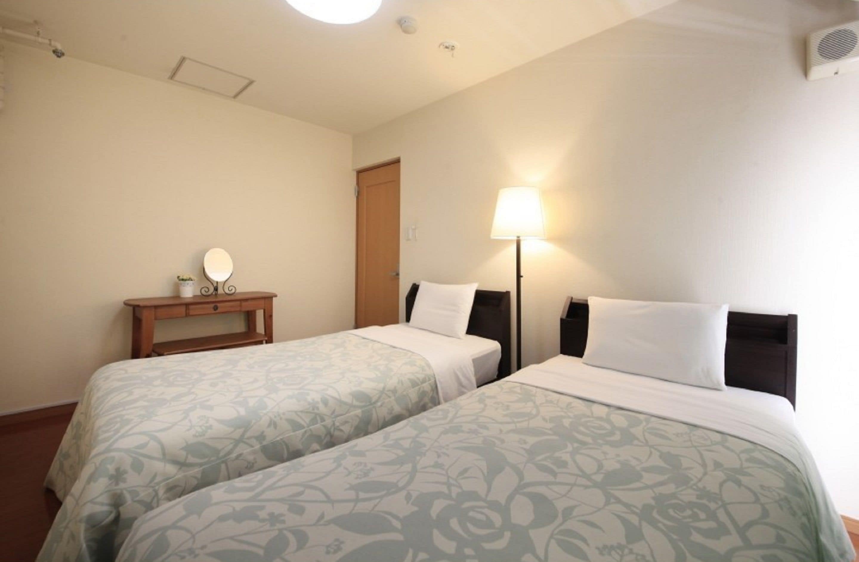 A hotel room with two beds, a bedside table with a lamp, a mirror, and a door.