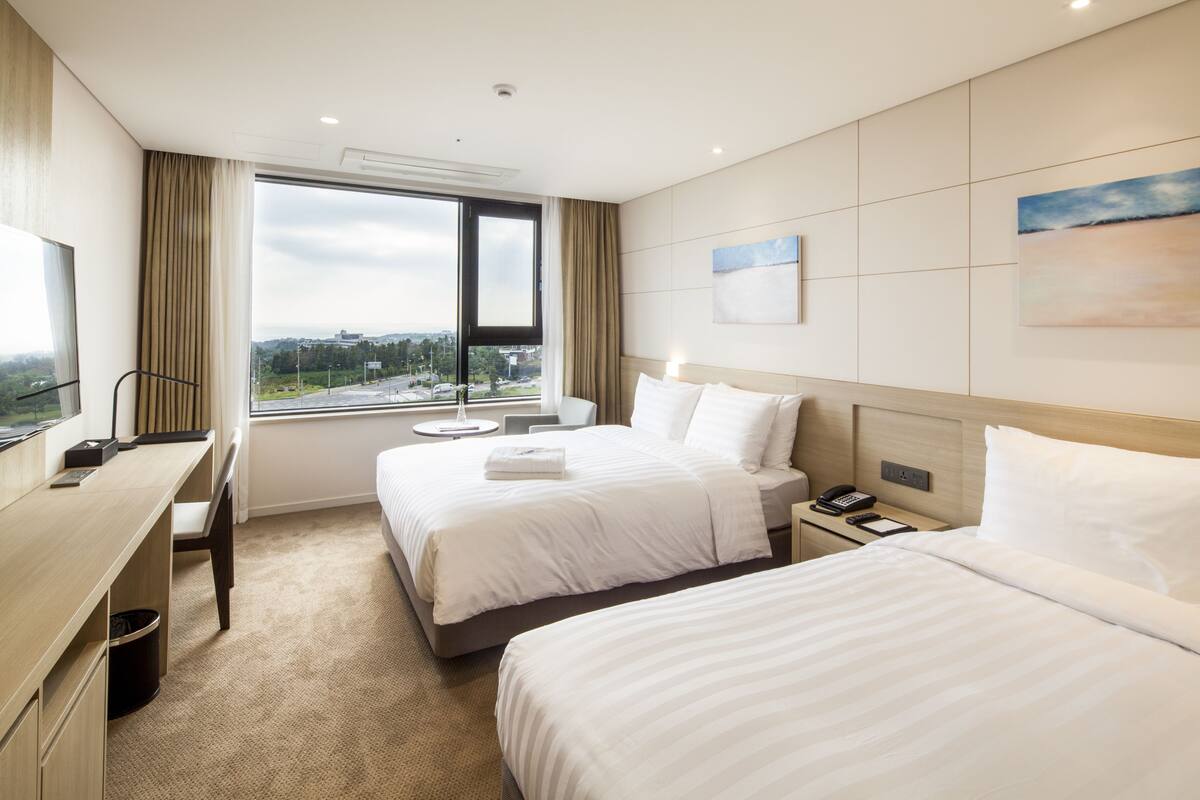 Een hotelkamer met twee bedden, een bureau, een stoel, een raam met uitzicht en een schilderij aan de wand.