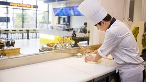 Un chef, vêtu d’une blouse et d’un chapeau blancs, travaille derrière un comptoir dans un café. Un panneau affichant le menu est visible en arrière-plan.