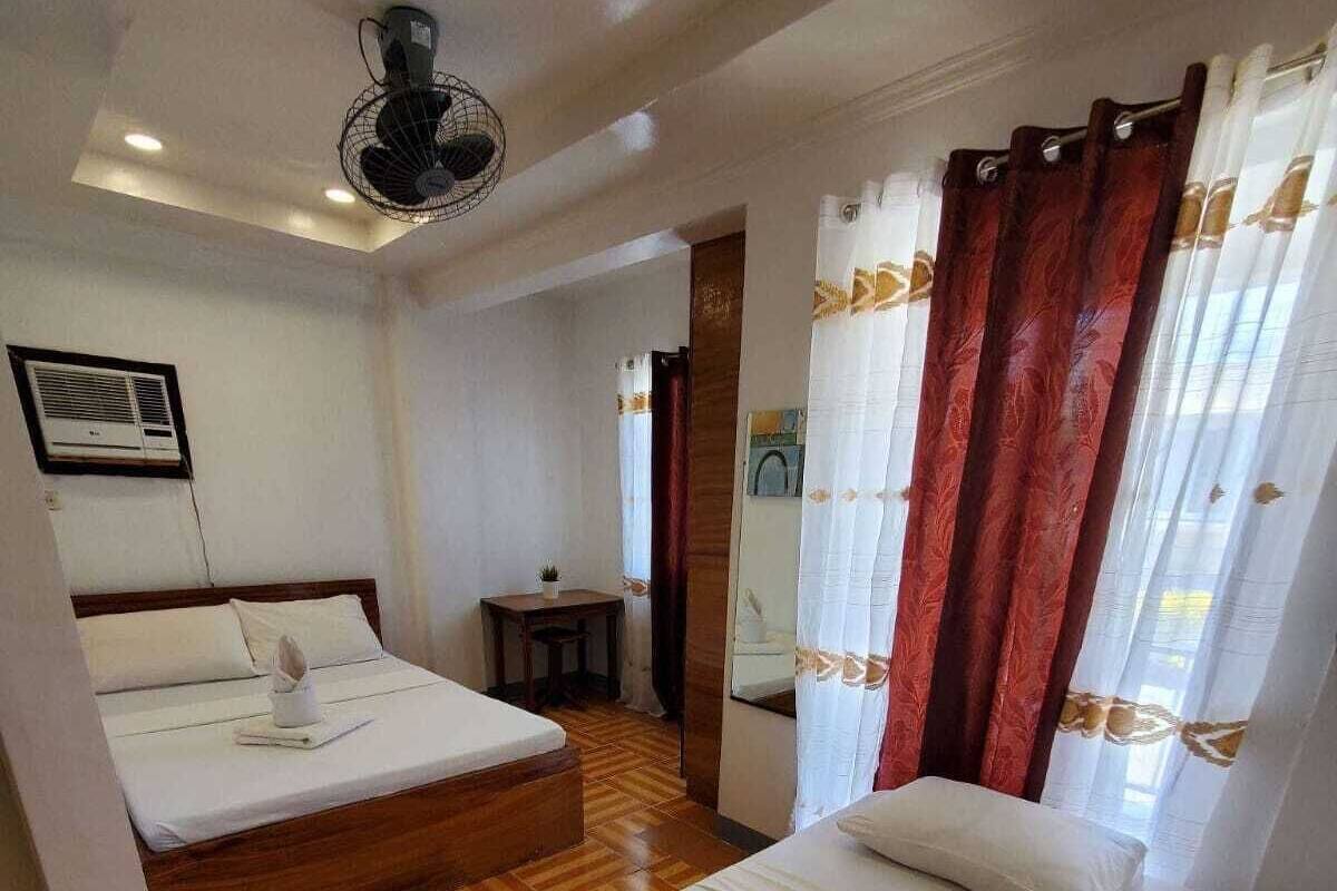 A hotel room with two beds, a small table, a fan, and a window with curtains.