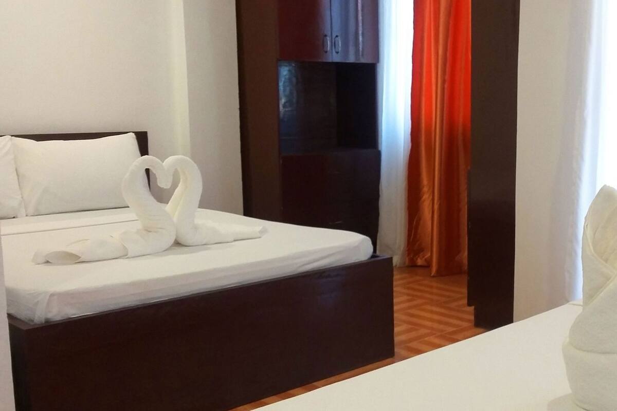A hotel room with a bed, a nightstand, a wooden floor, and a view of a room with a dark wall and orange curtains.
