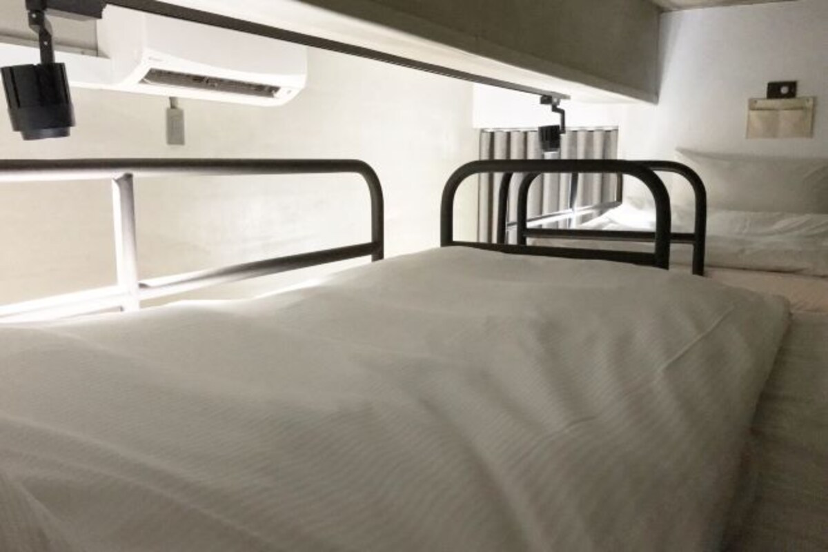 Ein Einzelbett mit Metallgestell, ein Nachttisch und eine an der Wand befestigte Lampe.