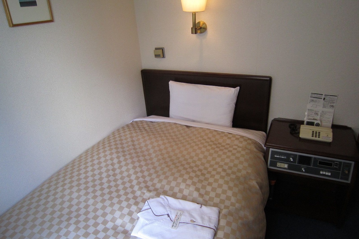 A hotel room with a checkered bedspread, a white pillow, a bedside table with a lamp, and a telephone.