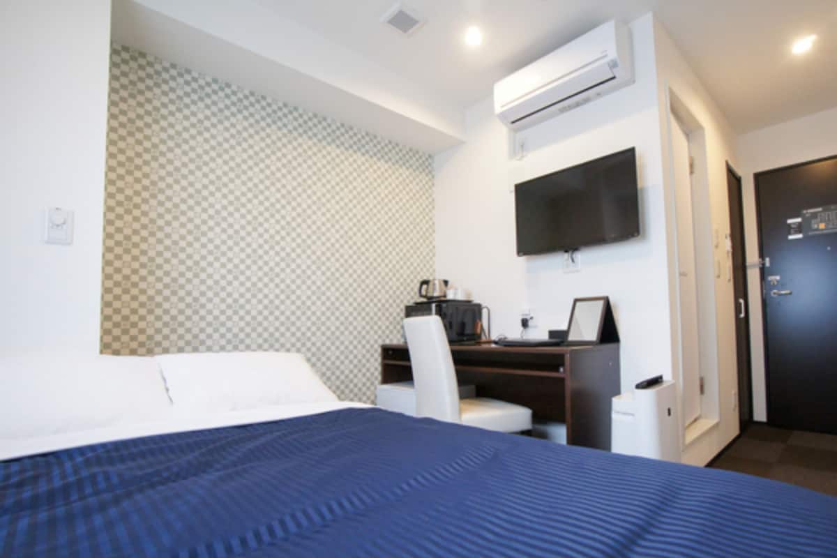 A hotel room with a bed, a desk with a computer, a chair, an air conditioning unit, a television, and a door.