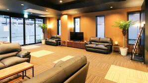 A modern lounge area with leather sofas, a flat-screen TV, and potted plants. - HOTEL LiVEMAX Minamihashimoto Ekimae (Sagamihara)