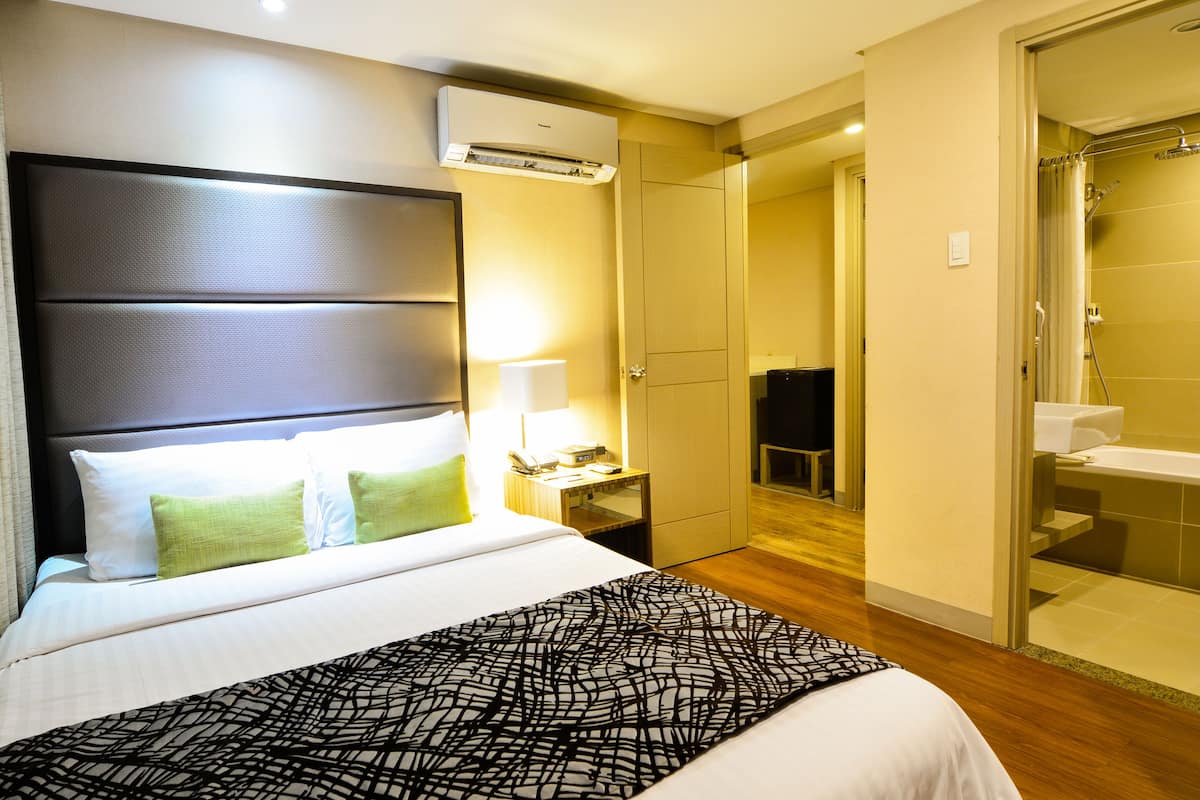 Habitación de hotel con una cama grande, mesita de noche, lámpara, aire acondicionado y vistas al baño.