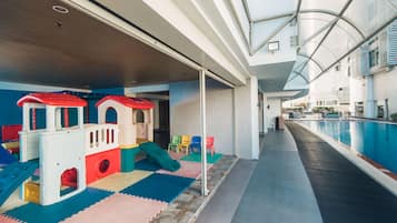 Children can play safely on the colorful play structures and soft mats near the pool, surrounded by a clear glass barrier.