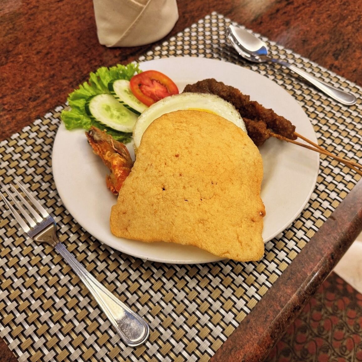 A plate with a fried item, a piece of meat, and vegetables on a woven placemat.
