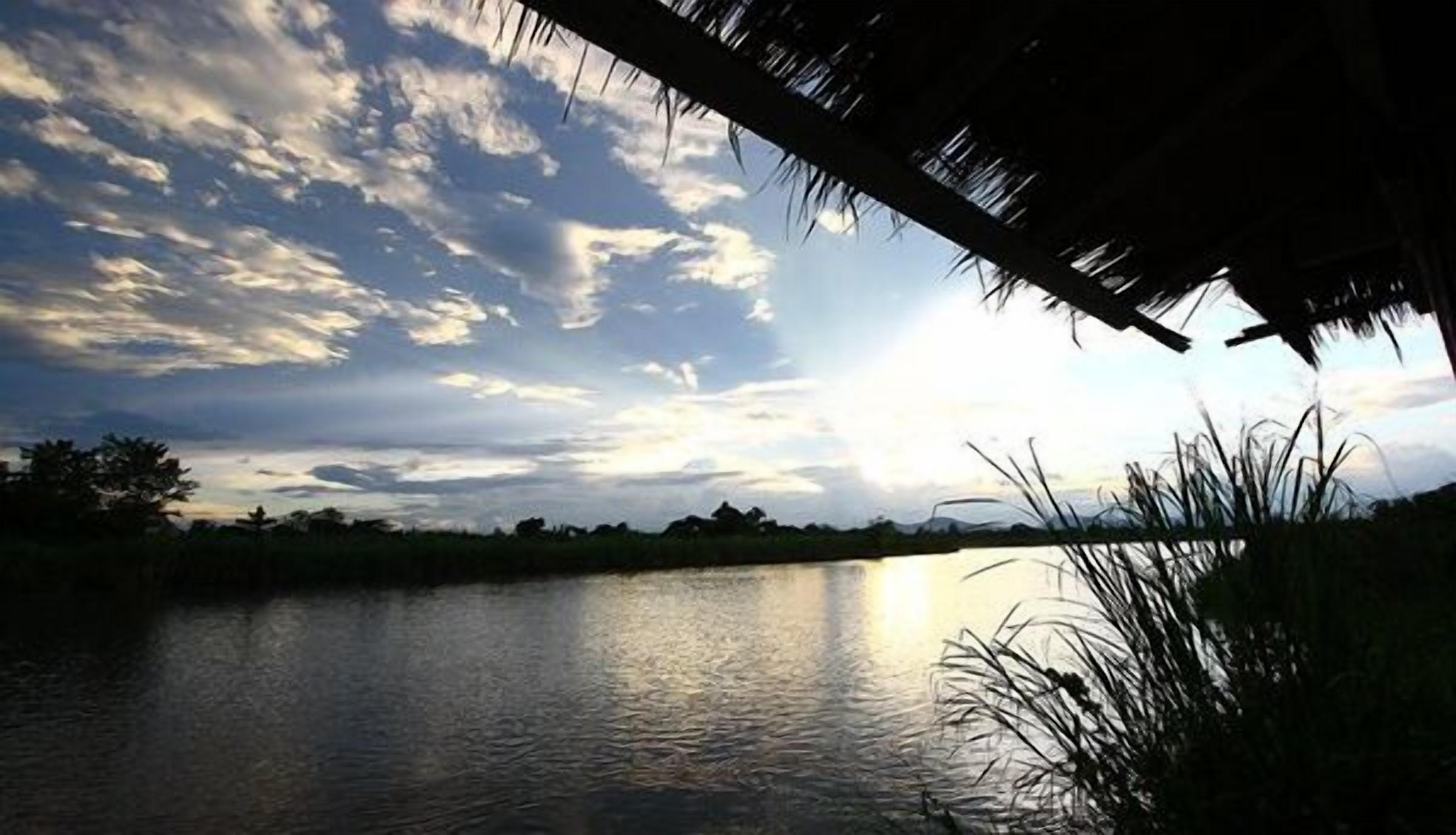 Una vista tranquila de un lago, con una estructura de techo de paja, agua calma y un cielo parcialmente nublado.