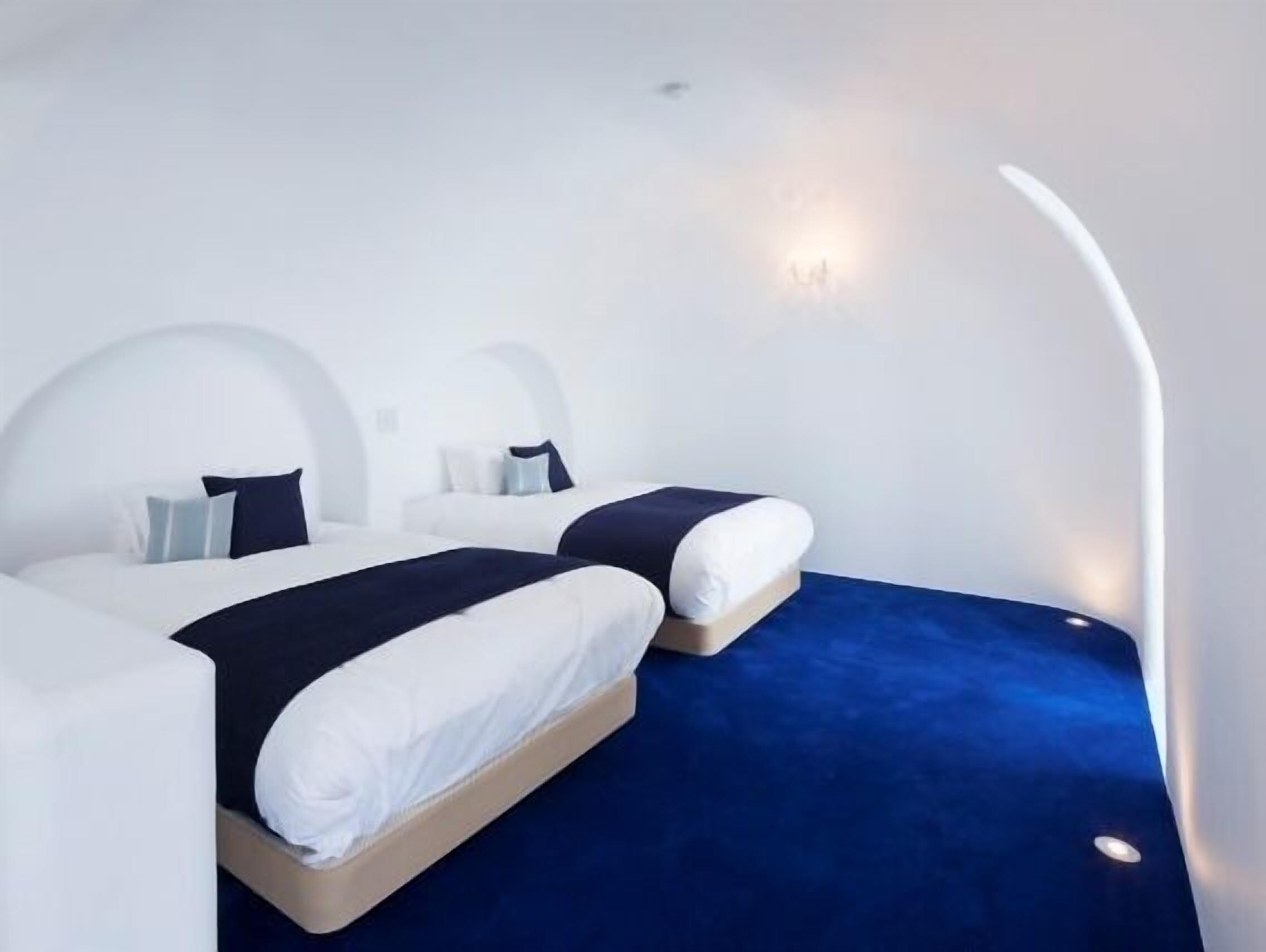 A modern hotel room with two beds, a blue carpet, and a curved wall design.