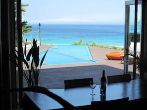 Blick von einem Zimmer mit Tisch und Stühlen auf einen Swimmingpool und den Ozean.