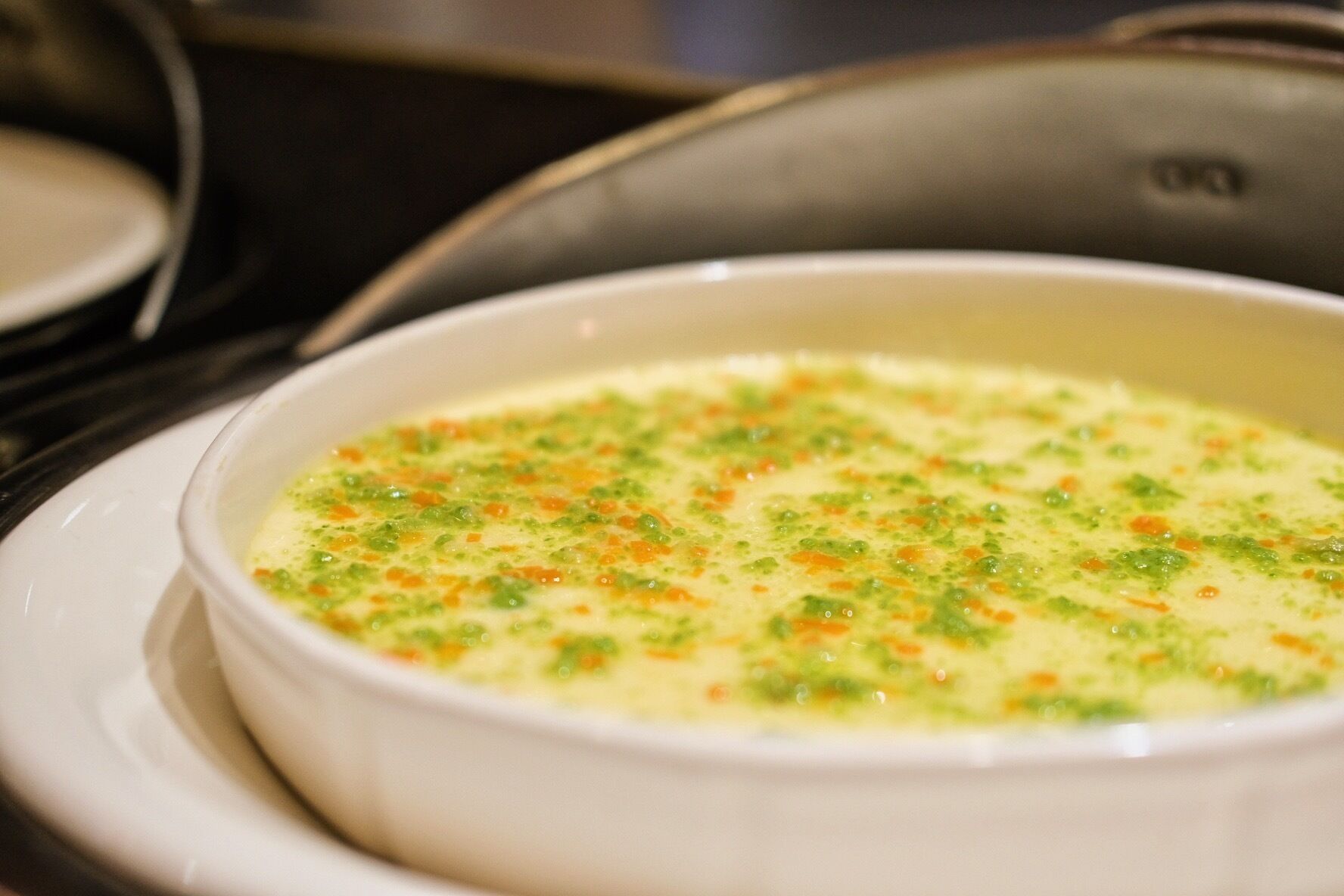 A white bowl of soup with green and orange specks on a white plate.